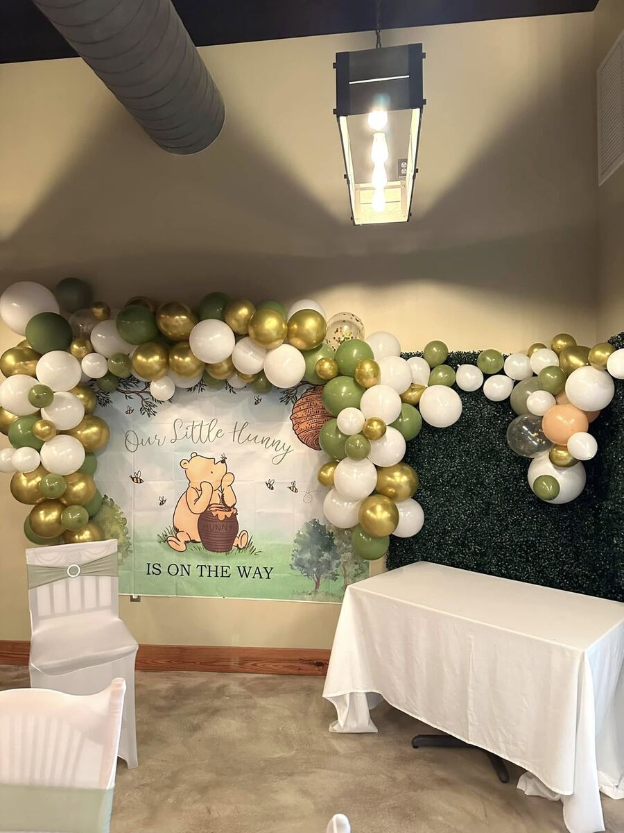 A Winnie-the-Pooh baby shower backdrop reading Our Little Hunny Is On The Way, framed by a sage, gold, and white balloon garland and a boxwood greenery wall.