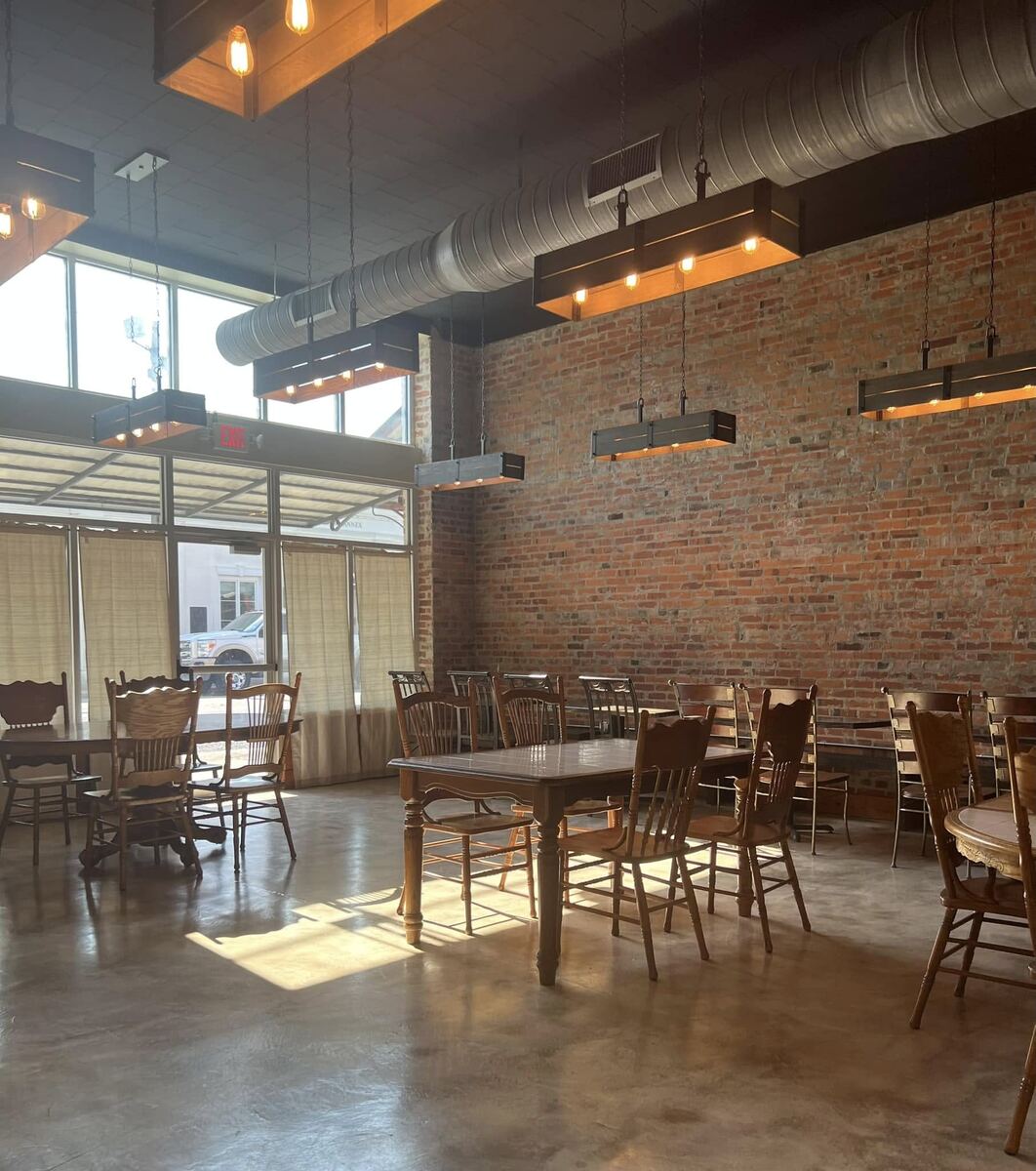 The open room at The Russo in daylight — a long exposed-brick wall, tall storefront windows, wooden tables and chairs, and industrial pendant lights on the high ceiling.