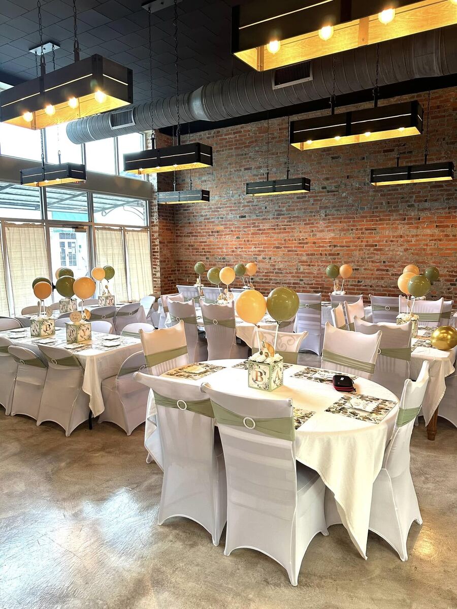 A baby shower set at The Russo, tables in white linens with sage sashes and balloon centerpieces against the exposed brick.