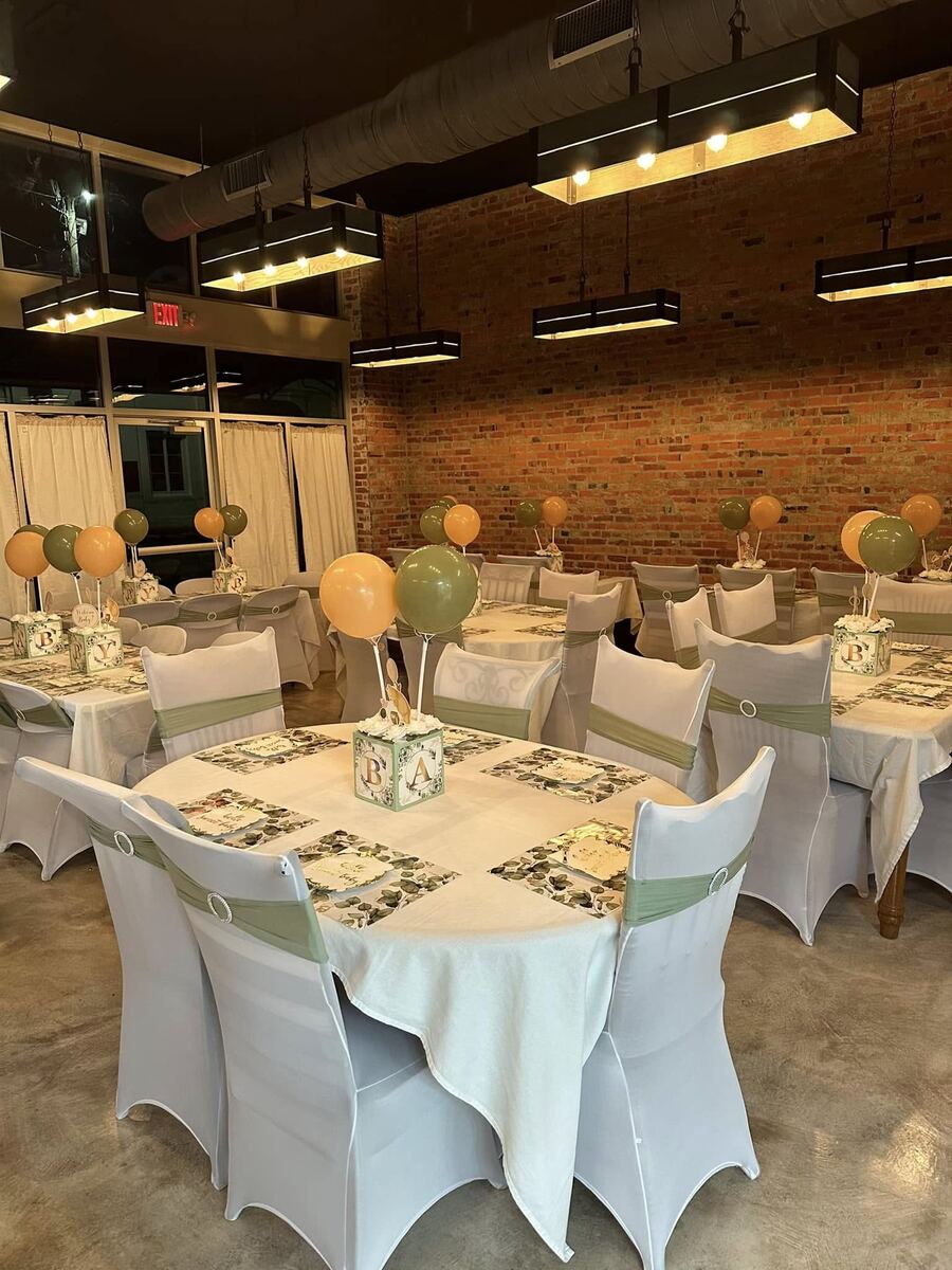 The Russo set for an evening baby shower — round tables in white covers with sage-ribboned chairs and balloon centerpieces glowing under warm pendant lights against the brick.