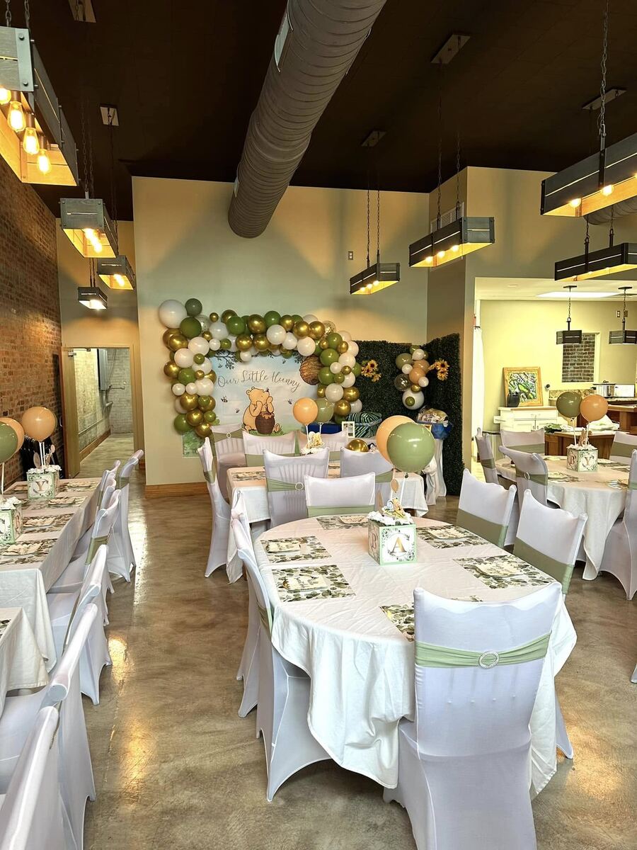 A styled baby shower filling the room at The Russo — tables in white linens with balloon centerpieces, a floral backdrop with sunflowers and a boxwood wall, and a view toward the back of the space.