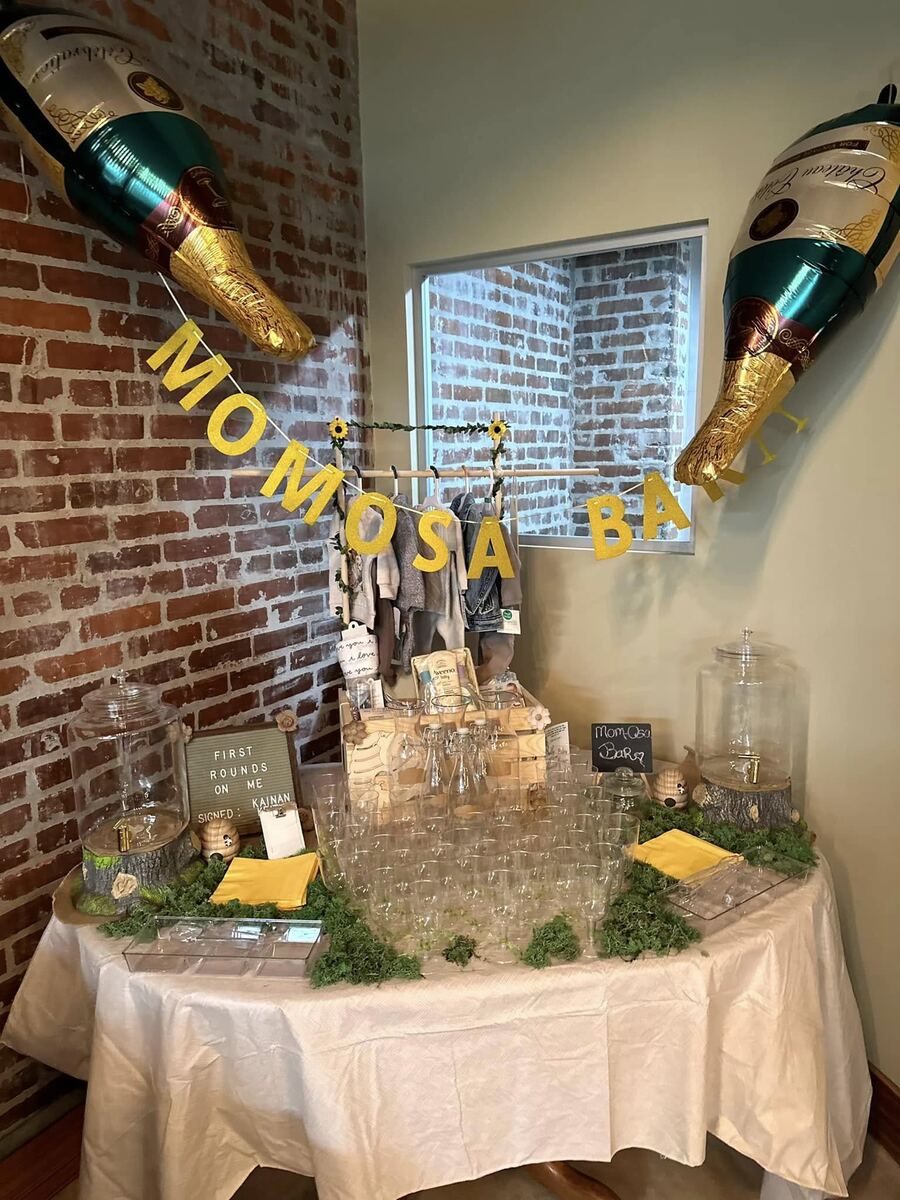 A styled Mom-osa Bar drink station at The Russo — glass beverage dispensers, rows of glasses, greenery, sunflowers, and giant champagne-bottle balloons against the exposed brick.