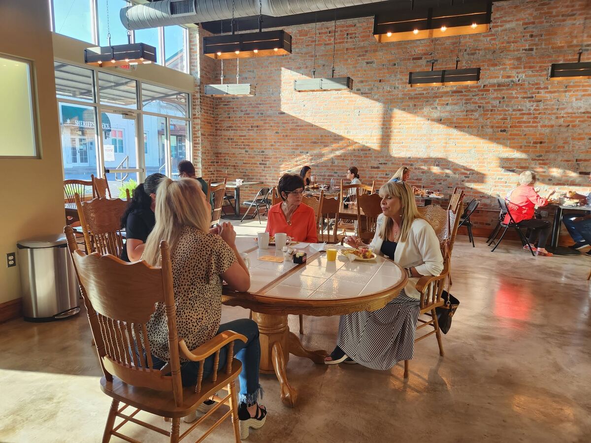 A gathering at The Russo — guests visiting over food at wooden tables, with downtown Abbeville visible through the tall front windows.