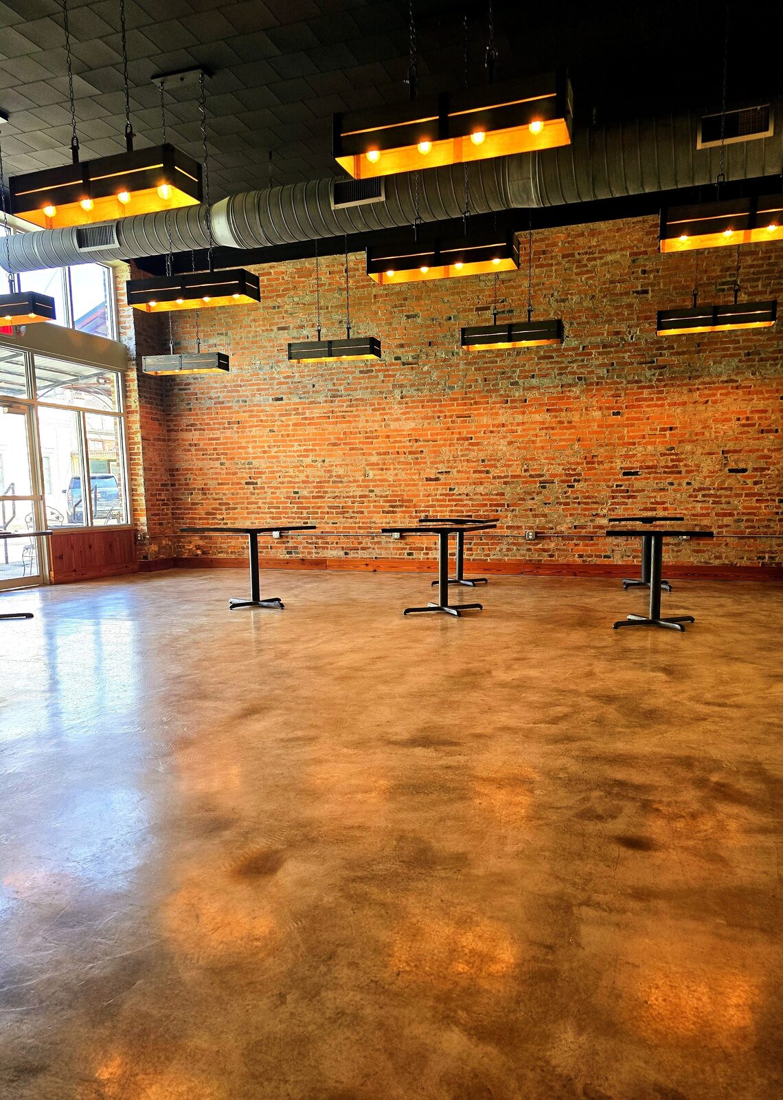 The main room at The Russo in warm light — a tall exposed-brick wall, industrial box pendant lights, a polished concrete floor, and pedestal tables, with downtown Abbeville through the storefront windows.