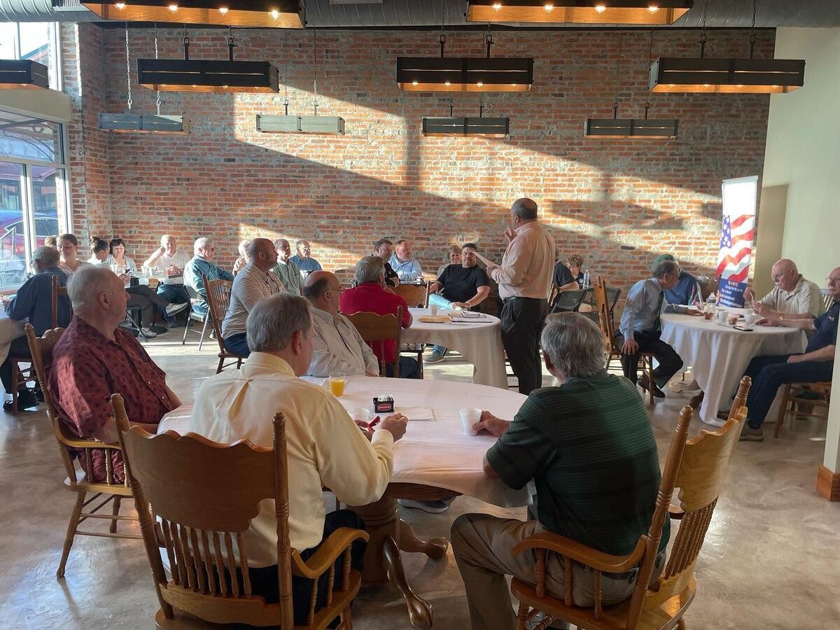 Neighbors gathered at round tables for a morning meeting inside The Russo, sunlight patterning the brick wall behind them.