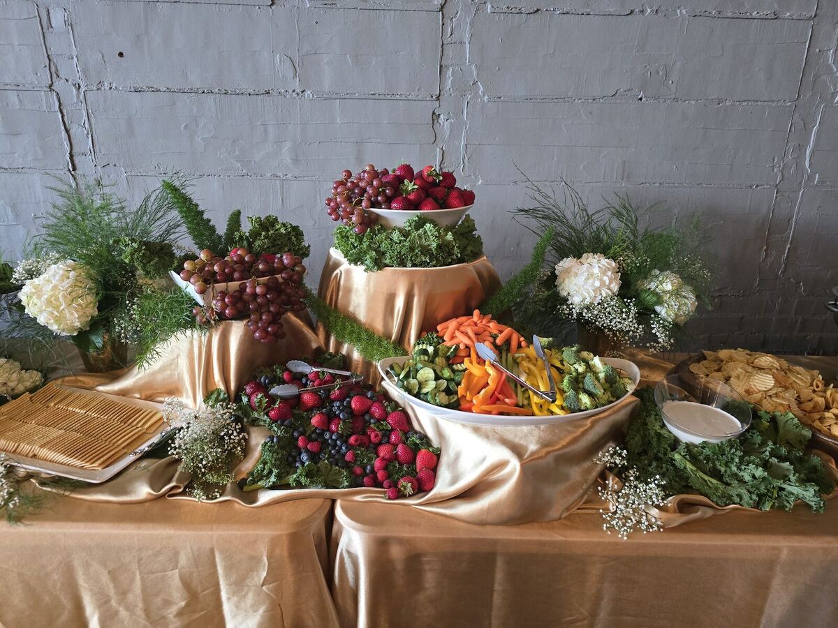 A grazing display by The Southern Spread on gold satin against a brick wall — tiered bowls of grapes and strawberries, a fresh vegetable platter, crackers, chips, and white hydrangea arrangements.