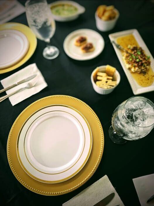 An elegant place setting on dark linen — a gold-rimmed charger and plate with appetizers and an iced glass of water.